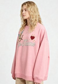 Jeune femme aux cheveux blonds ondulés portant un sweat-shirt rose oversize avec un cupidon, un cœur rouge et le texte "Je T'aime".