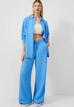 Kobieta ubrana w luźną niebieską koszulę zapinaną na guziki, na białym krótko ściętym topie i dopasowanych szerokich niebieskich spodniach, stoi z jedną ręką w kieszeni.