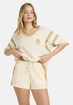 Off-white oversized t-shirt med grønne striber og en sol-logo; kombineret med cremefarvede shorts med orange sideaccenter og snøre i taljen.