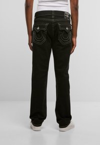 Jeans de mezclilla negra con costuras blancas, dos bolsillos traseros con un diseño curvado y un corte recto. Llevados con una camisa blanca y zapatillas.