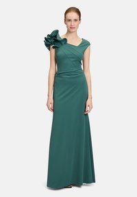 Robe de soirée teal en tissu lisse, présentant un design structuré, un décolleté asymétrique et un grand accent floral sur une épaule.