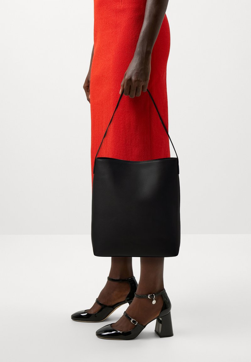 Sac en cuir noir avec une texture lisse et une seule bandoulière, tenu par une main à côté d'une silhouette vêtue d'une robe rouge, accentuée par des talons noirs.