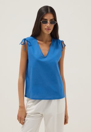 Donna con lunghi capelli scuri che indossa piccoli occhiali da sole rettangolari, un top blu senza maniche con scollo a V e dettagli annodati sulle spalle, e pantaloni bianchi.