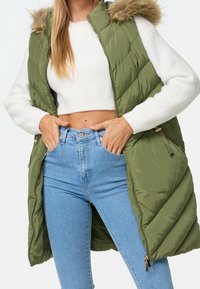 Vrouw in groene gewatteerde mouwloze jas met een bontgevoerde capuchon, witte cropped sweater en lichtblauwe high-waisted jeans, handen in de zakken.