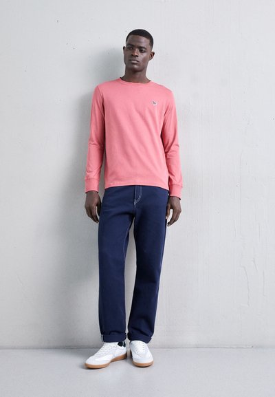 Long-sleeve coral shirt with a round neck, paired with navy wide-leg jeans and white sneakers with gum soles, standing against a gray wall.