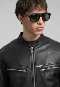 Zwarte leren jas met ritszakken, voorzien van metalen accenten en een opstaande kraag. Zwarte zonnebril met een logo op het montuur.
