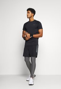T-shirt de sport noir, shorts noirs avec un motif sur le côté, leggings de compression gris et baskets blanches. Les bandeaux de poignet sont noirs avec le logo.