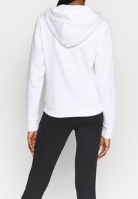 Sweat à capuche blanc avec une texture lisse, une coupe ajustée et des poignets côtelés. Porté avec un legging noir, montrant une tenue décontractée.