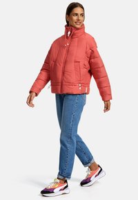 Koraal kleurige pufferjack met een hoge kraag, gestructureerd oppervlak, ritssluiting aan de voorkant, twee zijzakken en verstelbare trekkoorden. Draagt met lichtblauwe jeans en meerkleurige sneakers.