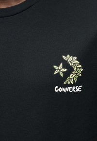Μαύρο βαμβακερό μπλουζάκι με σχέδιο πράσινου φύλλου και το κείμενο «CONVERSE» σε λευκό, τοποθετημένο στο πάνω αριστερό μέρος. Στρογγυλή λαιμόκοψη.