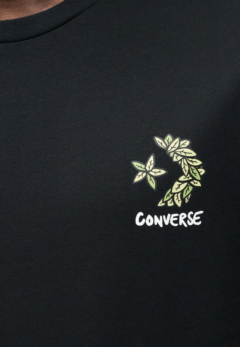 Maglietta nera in cotone con design a foglia verde e scritta "CONVERSE" in bianco, posizionata in alto a sinistra. Girocollo rotondo.