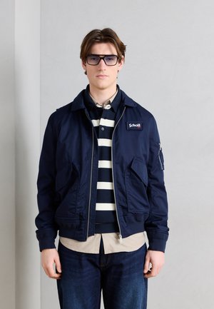 Jonge man met een marineblauw jack, een gestreepte polo, een beige overhemd, donkere spijkerbroek en getinte zonnebril tegen een effen lichtgrijze achtergrond.