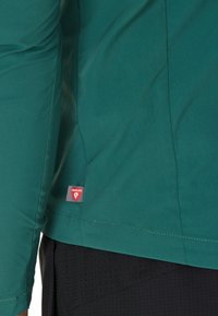 Maglietta a maniche lunghe color turchese con una texture liscia, dotata di un'etichetta del marchio rossa a contrasto. La maglietta ha una vestibilità sagomata e dettagli di cucitura puliti.