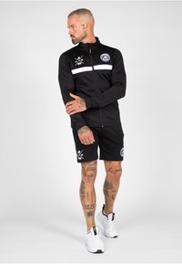 Zwarte zip-up jack met witte streep en logo, gecombineerd met zwarte shorts met logo en grafische print. Model draagt witte sportschoenen.