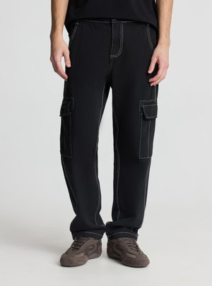 Pantaloni cargo da uomo neri con cuciture a contrasto bianche e tasche con patta, abbinati a sneakers marroni, in piedi su uno sfondo neutro.