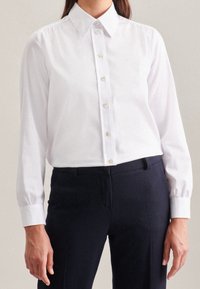 Vit långärmad skjorta med knappar, klassisk krage, knäppning fram och samlade ärmkanter. Matchas med mörka byxor.