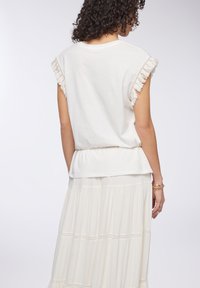 Camicia bianca di cotone con dettagli di frange sulle maniche corte, vita arricciata, vista da dietro, abbinata a una gonna fluida color crema.