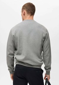 Sudadera gris claro de manga larga, con cuello redondo, puños y cintura acanalados, combinada con pantalones negros, vista desde la parte posterior.