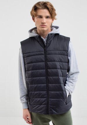 Czarna, pikowana kamizelka z zamkiem z przodu, z wysokim kołnierzem i bez rękawów. Noszona na szarym hoodie i zestawiona z zielonymi spodniami.