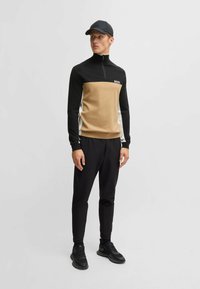 Schwarzer und beigefarbener Sportpullover mit Reißverschluss und "BOSS"-Logo, kombiniert mit schwarzen Sporthosen und schwarzen Sneakers, getragen mit einer passenden Mütze.