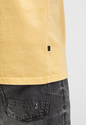 Gele zoom van overhemd met klein zwart label en versleten zwarte denim spijkerbroekzak met witte geborduurde letter J en verweerd detail.