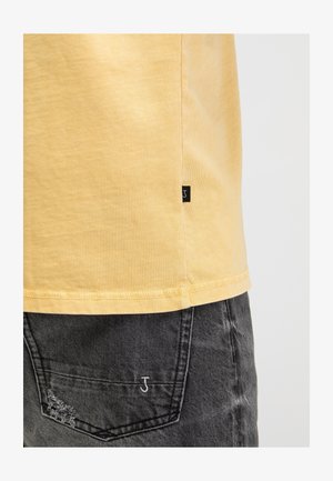 Gelber Hemdsaum mit kleinem schwarzem Etikett und abgenutzter schwarzer Jeans-Tasche mit weiß besticktem Buchstaben J und Used-Detail.
