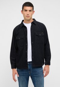 Schwarze Cord-Hemdjacke mit zwei Brusttaschen, Knopfverschluss und lockerem Schnitt über einem weißen T-Shirt, kombiniert mit blauen Jeans.