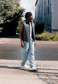Black quilted sleeveless jacket over a white shirt, light blue baggy jeans, and white sneakers. Standing outdoors near a building.