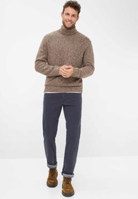 Maglione a collo alto lavorato a maglia marrone con motivo testurizzato, abbinato a pantaloni blu scuro e stivali stringati marroni, in piedi davanti a uno sfondo chiaro.