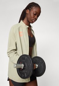 Light olive athletic jacket with a high collar, orange graphic text, paired with a black sports bra and holding a black weight.