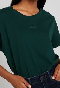 Modelka v tmavě zeleném tričku s jemným logem, ruce na bocích, v kombinaci s tmavými džínami, předvádějící casual outfit.