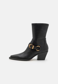 Bota de cuero negro hasta el tobillo con punta afilada y una correa texturizada con un hebilla dorada. El tacón es de forma cuadrada y de altura media.