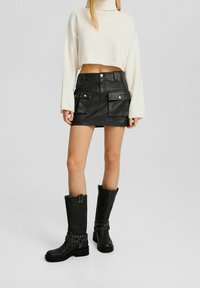 Krämfärgad kortärmad tröja, svart läder mini mörk kjol med framfickor, och knähöga svarta stövlar med metalliska accenter och texturerad finish.