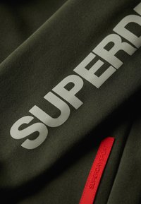 Tessuto verde oliva con una consistenza morbida, presenta una grande scritta bianca che recita "SUPERDRY" e un piccolo etichetta rossa con la dicitura "SUPERDRY SPORT".