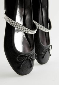 Schwarze Ballettschuhe aus Stoff mit einem glitzernden silbernen Strassriemen, dekorativem Schleifen und glattem, glänzendem Innenraum.