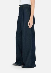 Pantaloni in denim a gamba larga in lavaggio blu scuro, con vita alta, pinces e cuciture a contrasto. L'orlo tocca le scarpe, evidenziando una vestibilità rilassata.