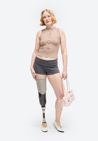 Woman with short blonde hair wearing a sleeveless beige top, gray shorts, white shoe, and a prosthetic left leg, holding a light pink handbag.