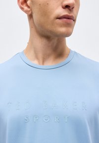 Κοντινό πλάνο ενός ατόμου που φοράει ανοιχτό μπλε πουκάμισο με ανάγλυφο κείμενο «TED BAKER SPORT» στο στήθος, στραμμένο ελαφρώς προς τα δεξιά.