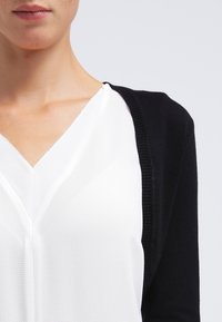 Frau in einer weißen V-Ausschnitt-Bluse unter einem schwarzen Cardigan, vom Kinn bis zur Mitte des Oberkörpers vor einem einfarbigen Hintergrund abgebildet.