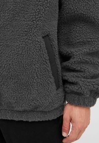 Mørkegrå fleecepullover med en blød, tekstureret overflade, der har en sort stoflomme på venstre side og elastiske manchetter.