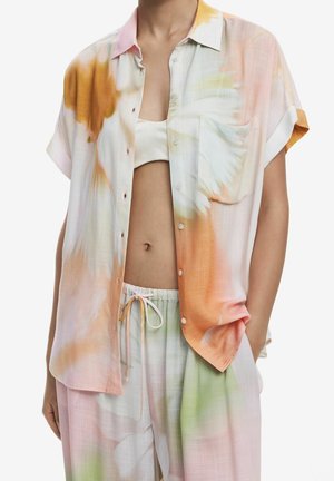 Person iført pastel-farvet tie-dye kortærmet skjorte med knapper over en hvid crop top og matchende bukser med snøre, hænderne i lommerne.