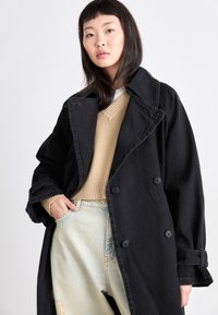 Manteau trench noir en coton avec un design à double boutonnage, grand col et poignets retroussés. Associé à un pull beige et un jean déchiré bleu clair.