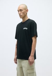 Čierne tričko s okrúhlym výstrihom, krátkymi rukávmi a prednou potlačou loga. Hladká bavlnená textúra, kombinované s khaki cargo nohavicami.