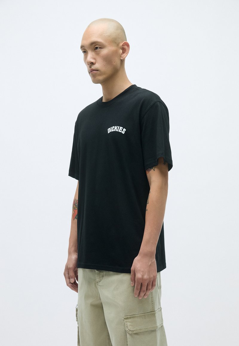 Schwarzes T-Shirt mit Rundhals, kurzen Ärmeln und Drucklogo auf der Vorderseite. Glatte Baumwolltextur, kombiniert mit khakifarbenen Cargo-Hosen.