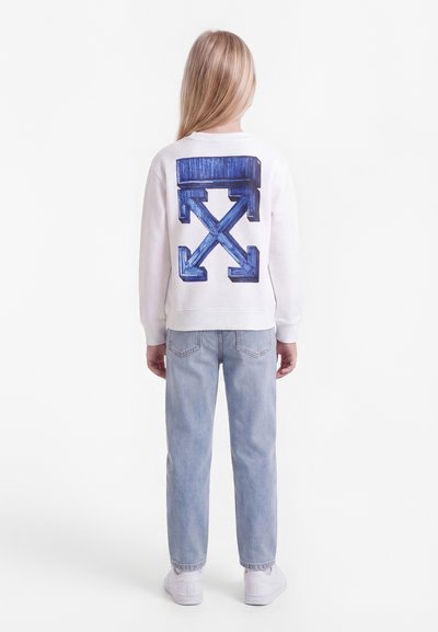 Παιδί με μακριά ξανθά μαλλιά που στέκεται, φοράει λευκό φούτερ με μπλε βέλος στο πίσω μέρος, ανοιχτό μπλε τζιν και λευκά αθλητικά παπούτσια.