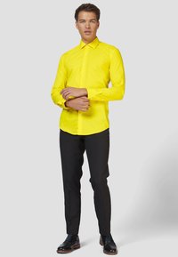 Camicia da uomo a maniche lunghe gialla con design a bottoni, abbinata a pantaloni neri su misura e scarpe nere, su uno sfondo neutro.