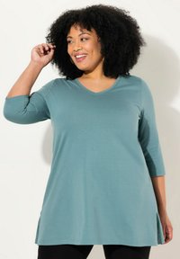 Tunique verde acqua con scollo a V e maniche a tre quarti, con spacchi laterali e una texture liscia, abbinata a leggings neri.
