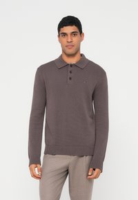 Mann trägt einen braunen, langärmligen Strick-Polopullover und hellbeige Hosen, steht vor einem schlichten weißen Hintergrund.