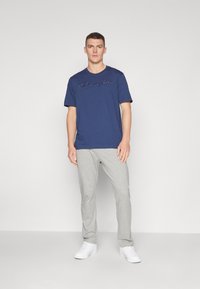 Dunkelblaues T-Shirt mit kurzärmeligem Schnitt und gesticktem "Champion"-Logo, kombiniert mit hellgrauen Sweatpants und weißen Sneakers.