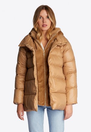 Beige Pufferjacke mit gestepptem Design, Druckknöpfen und einem weichen Innenfutter. Verfügt über eine Kapuze und einen Reißverschluss. Getragen über einer inneren Schicht in ähnlichem Farbton.